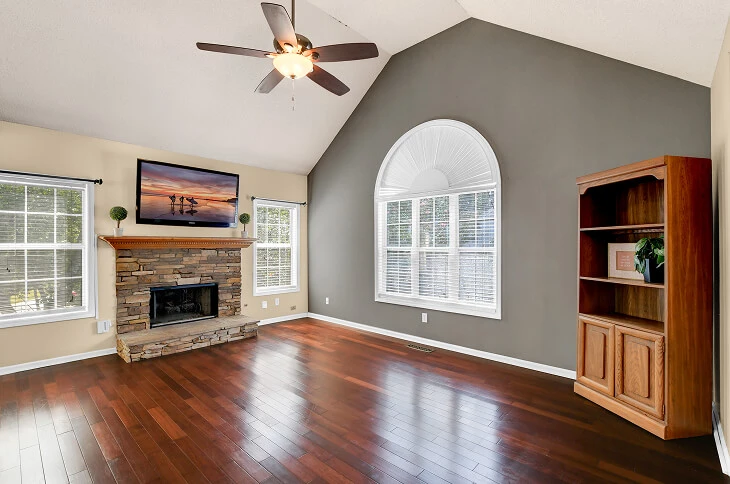 Before photo of empty living room with stone fireplace, vaulted ceiling, and large arched window, prior to Blue Dot Photos HDR enhancement.