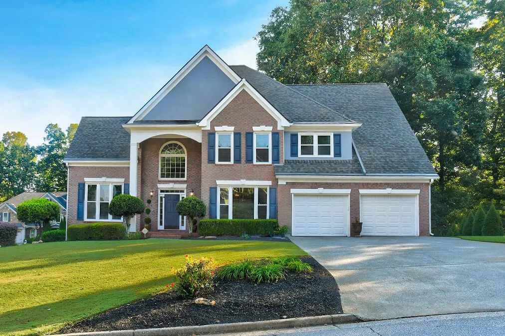 Modern two-story brick house with white garage doors, manicured lawn, and large windows surrounded by trees on a sunny day Modern two-story brick house with white garage doors, manicured lawn, and large windows surrounded by trees on a sunny day