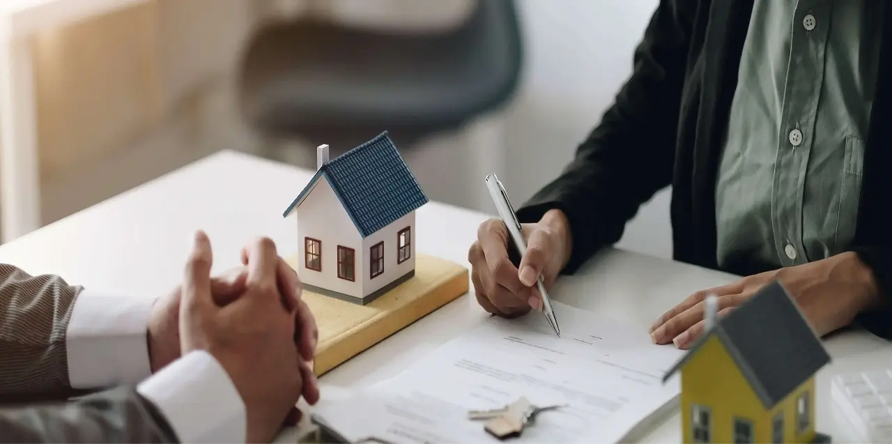 A person in a white shirt holding a small toy house model on a desk, symbolizing real estate ownership and property investment.