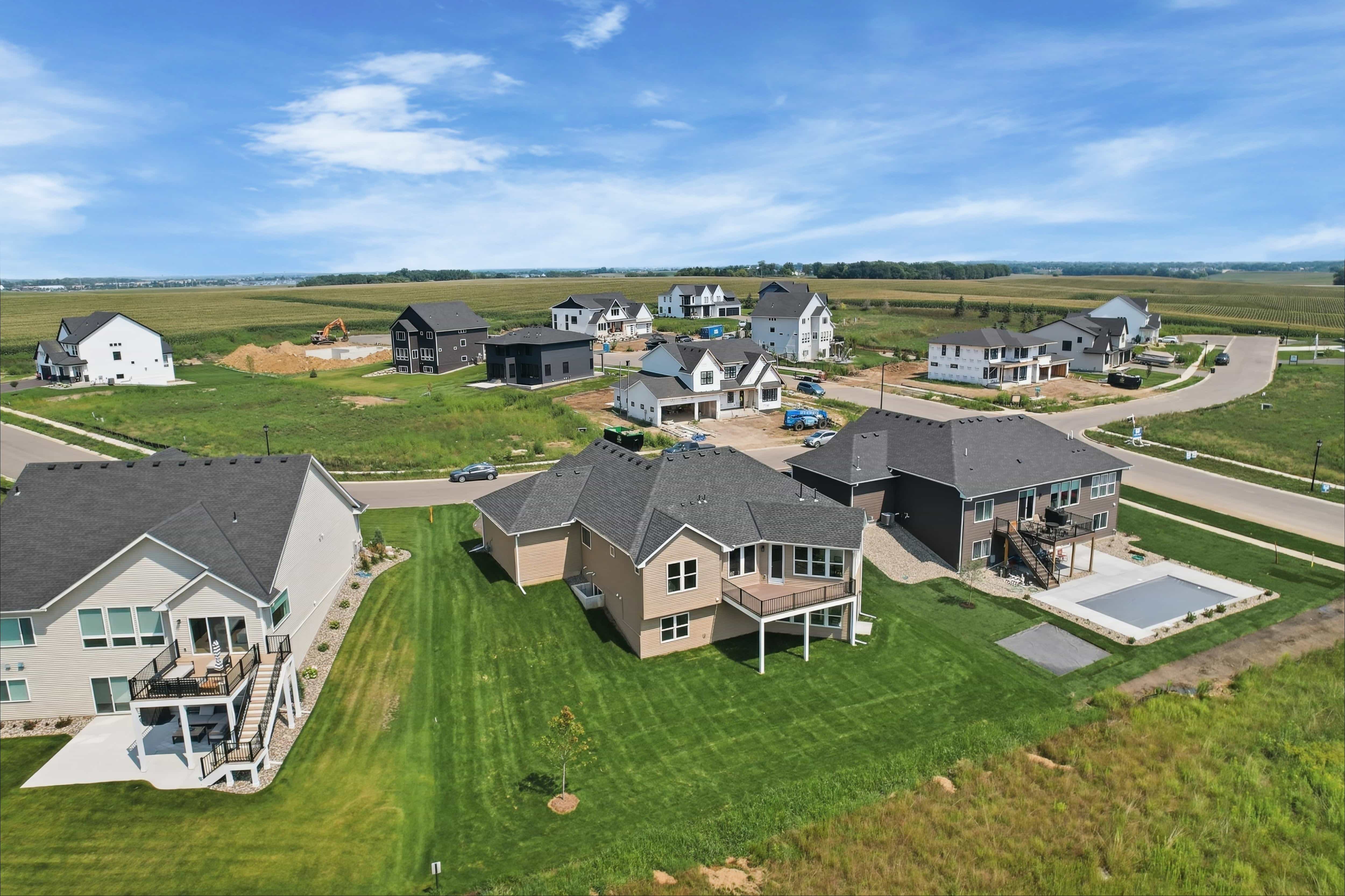 Aerial drone photo of a new construction neighborhood with two-story homes, backyard decks, and an in-ground pool, bordered by cornfields.