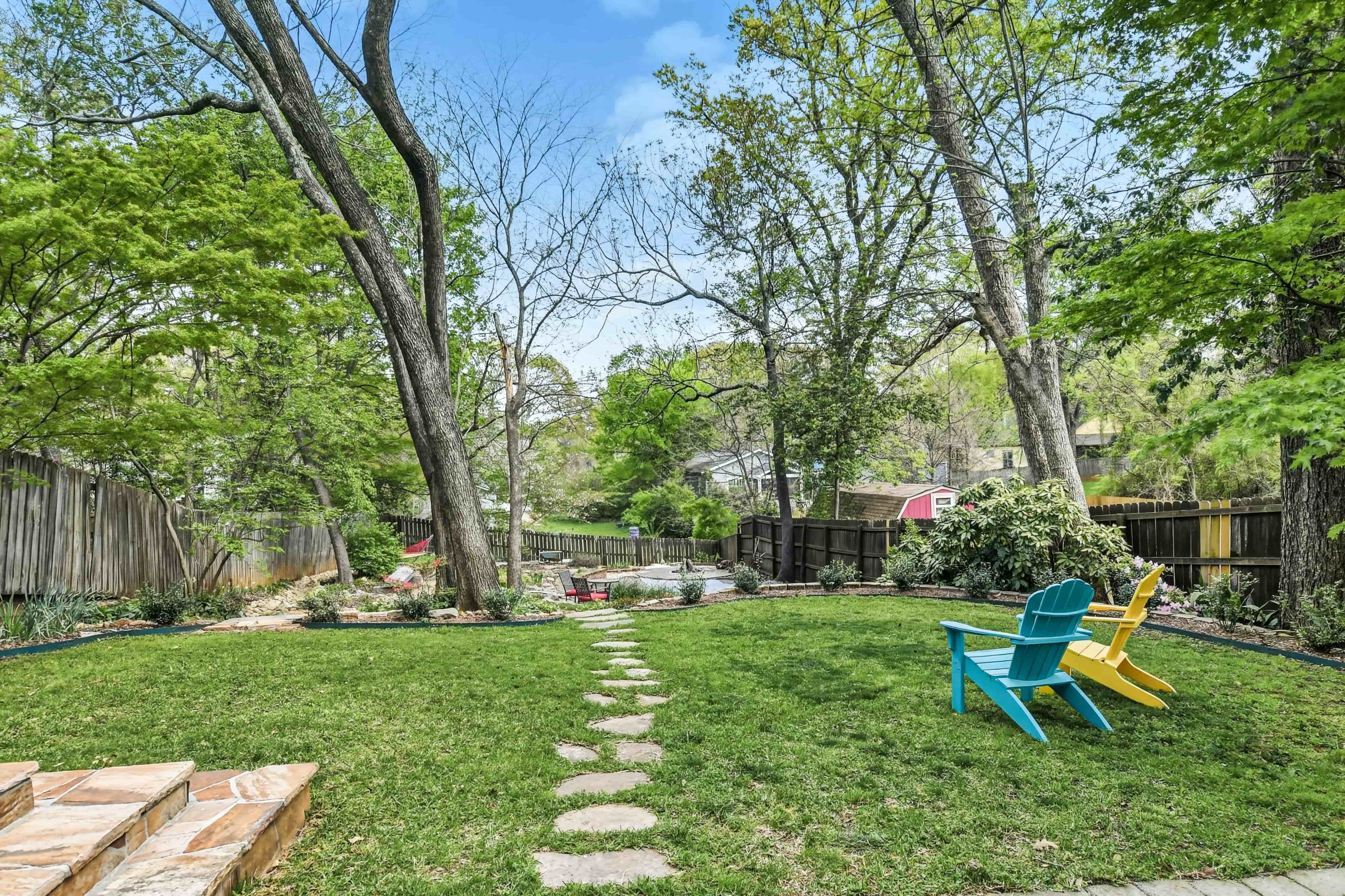 Fenced backyard with a green lawn, stepping-stone path, Adirondack chairs, and mature shade trees